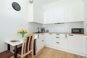 Una cucina compatta con mobili bianchi, un forno a microonde, un bollitore e una zona pranzo con una sedia e un tavolo.