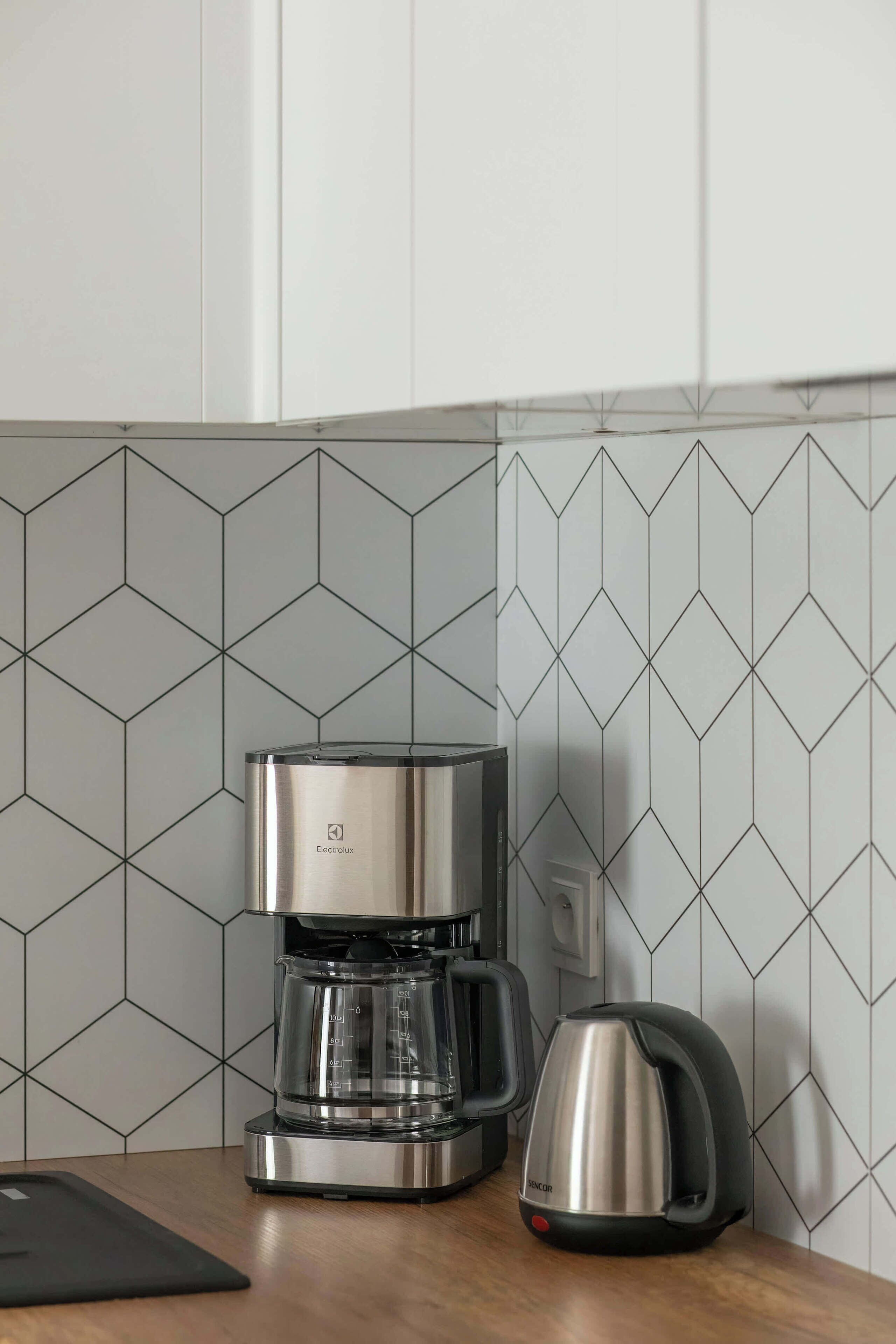 Una cocina con cafetera y hervidor sobre la encimera, un tapete negro y un salpicadero de azulejos.