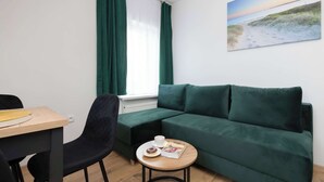 A room with a green sofa, a small round table with a cup and a book, a chair, and a window with green curtains.