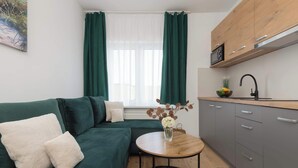 A modern living room with a green sofa, a small round table with a vase, and a kitchen area with grey cabinets and a black faucet.