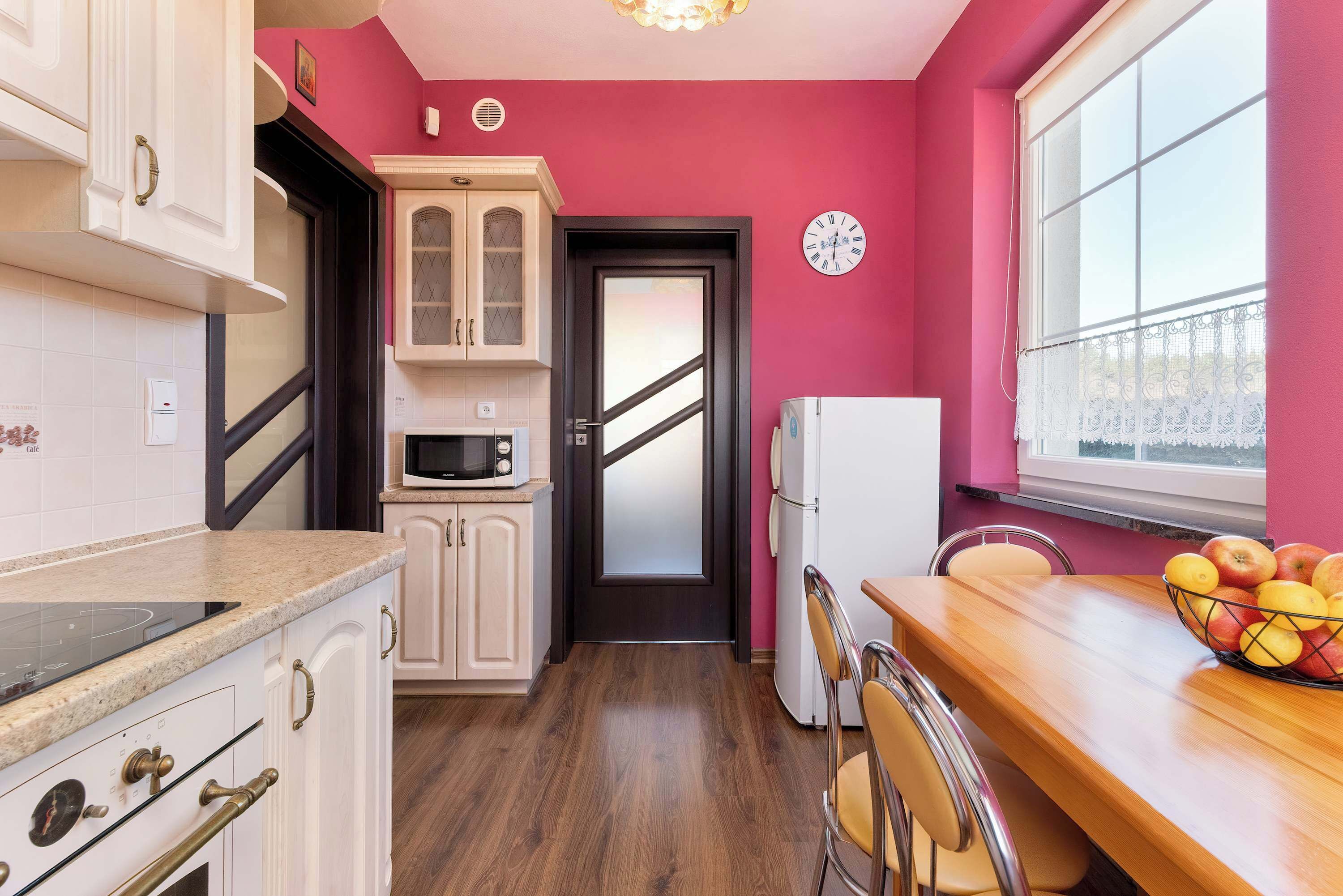 Una cocina con armarios blancos, un microondas, un refrigerador, una mesa de comedor de madera con sillas y un frutero.