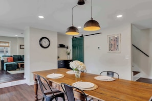 A dining area with a wooden table set for four, two pendant lights, a clock, and a green door in the background. - Downtown Saratoga Home w/ Yard & Patio (Saratoga Springs)