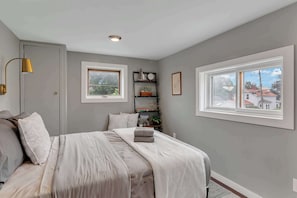 A bedroom with a bed, two windows, a shelf with books and a clock, and a lamp. - Downtown Saratoga Home w/ Yard & Patio (Saratoga Springs)