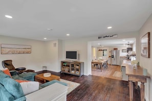 A spacious living room with a large sofa, a coffee table, and a TV on a cabinet. There is a dining area with a table and chairs, and a kitchen in the background. - Downtown Saratoga Home w/ Yard & Patio (Saratoga Springs)