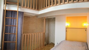 A room with a wooden ladder, a bed with a light-colored mattress, a wall with a textured finish, and a wall-mounted light fixture.