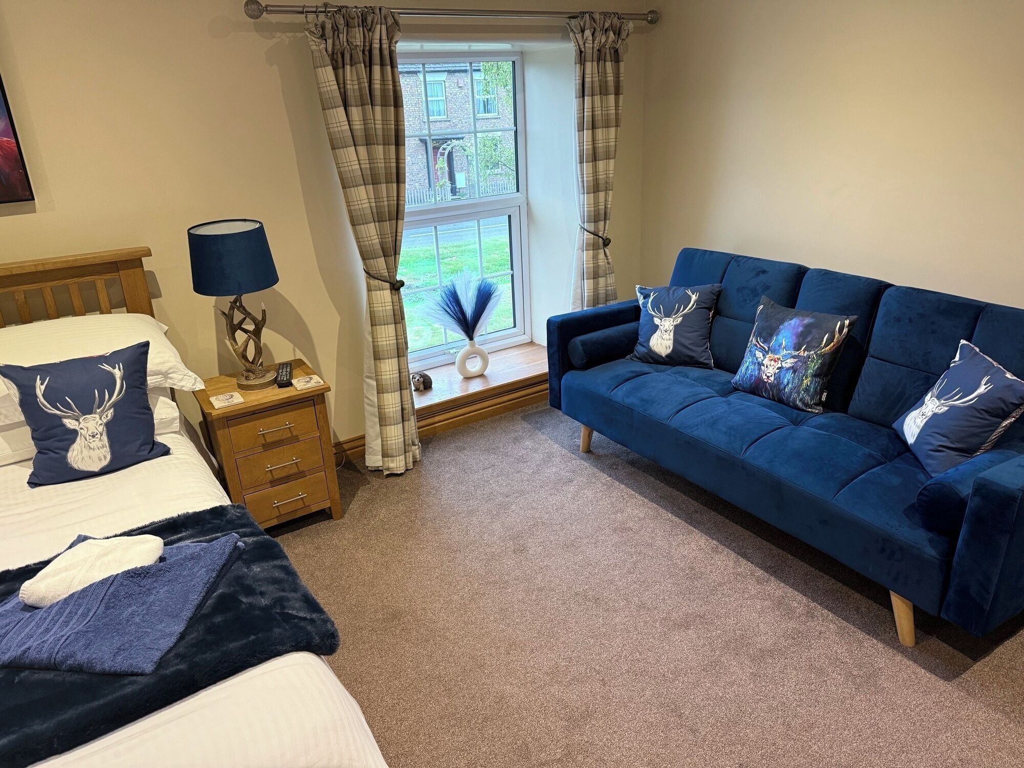 A bedroom with a bed, a nightstand, a lamp, a blue sofa with deer-themed pillows, and a window with curtains.