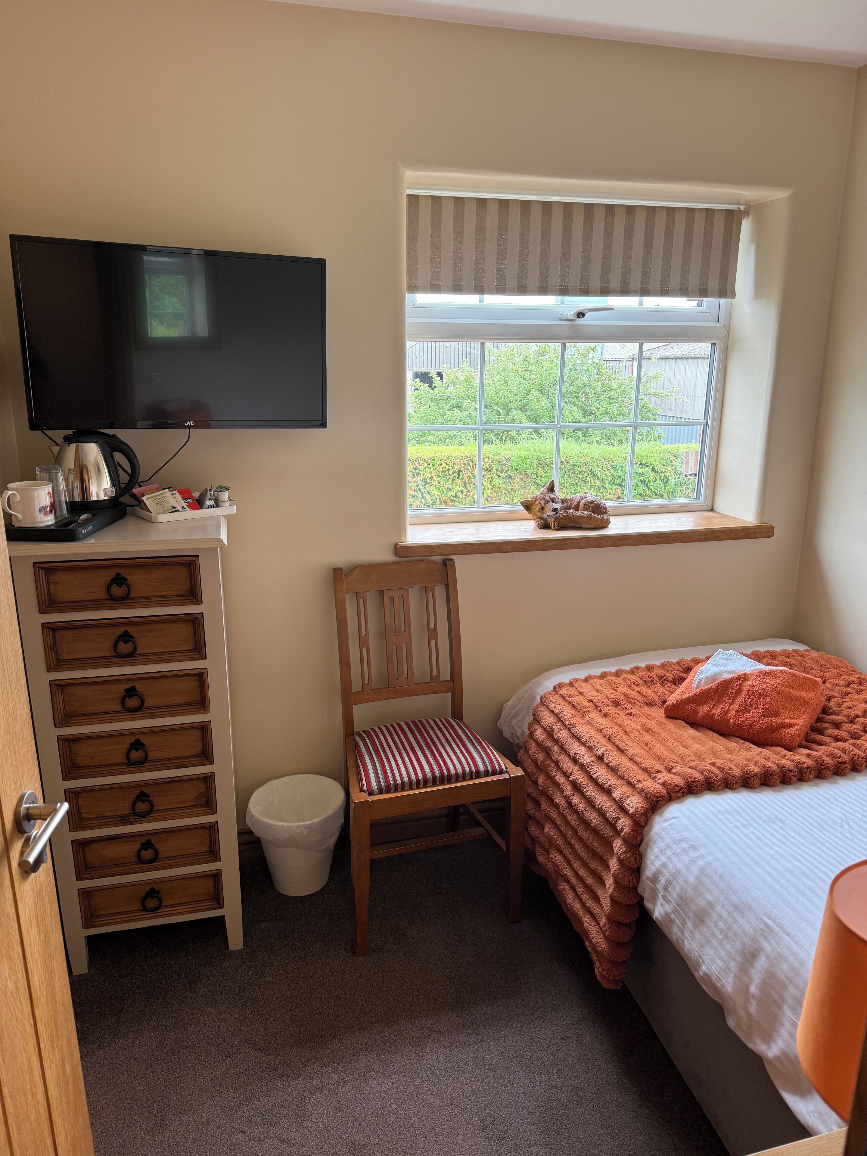 Ein Schlafzimmer mit einem Bett, einem Sessel, einem Fernseher, einem Fenster mit Blick auf Grünflächen und einem kleinen Tisch mit Wasserkocher und Tassen.