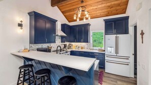 Une cuisine moderne avec des meubles de couleur bleu foncé, un îlot central blanc doté de tabourets, un réfrigérateur en acier inoxydable et un plafond en bois.