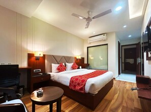 A hotel room with a bed, bedside tables, a desk, a chair, a round table, a ceiling fan, and an air conditioning unit. - Hotel The crystal Park  (Gwalior)