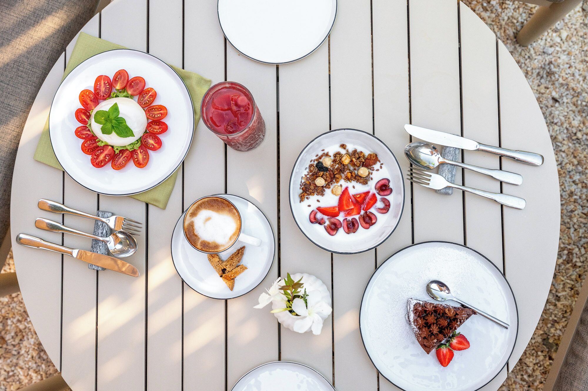 A round table with various dishes including a plate of sliced tomatoes, a bowl of yogurt with fruit, a slice of cake, and a cup of coffee.