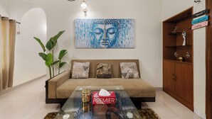 Uma sala de estar com um sofá, uma mesa de centro com um estojo de lenços, uma pintura de Buda, uma planta em vaso e um armário de madeira com prateleiras.