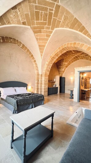 Una habitación amplia con cama, una mesa y zona de estar. Destacan paredes de ladrillo visto y un techo alto.
