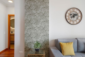A room with a patterned wallpaper, a glass table with a plant, a grey sofa with a yellow pillow, and a wall clock.