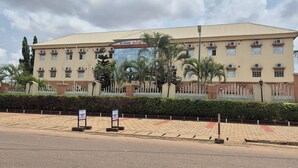 A large, multi-story building with a flat roof and multiple windows, surrounded by a fence and palm trees. - Al-Ihsan Metro Hotel (Kaduna)
