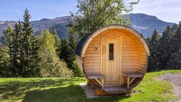 Une structure de sauna en bois, dotée d’une porte et d’un escalier, entourée d’herbe et d’arbres, avec des montagnes en arrière-plan.