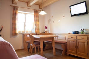 Ein gemütliches Zimmer mit Holztisch, einer Bank, einem an der Wand befestigten Fernseher und einem Fenster mit Vorhängen.