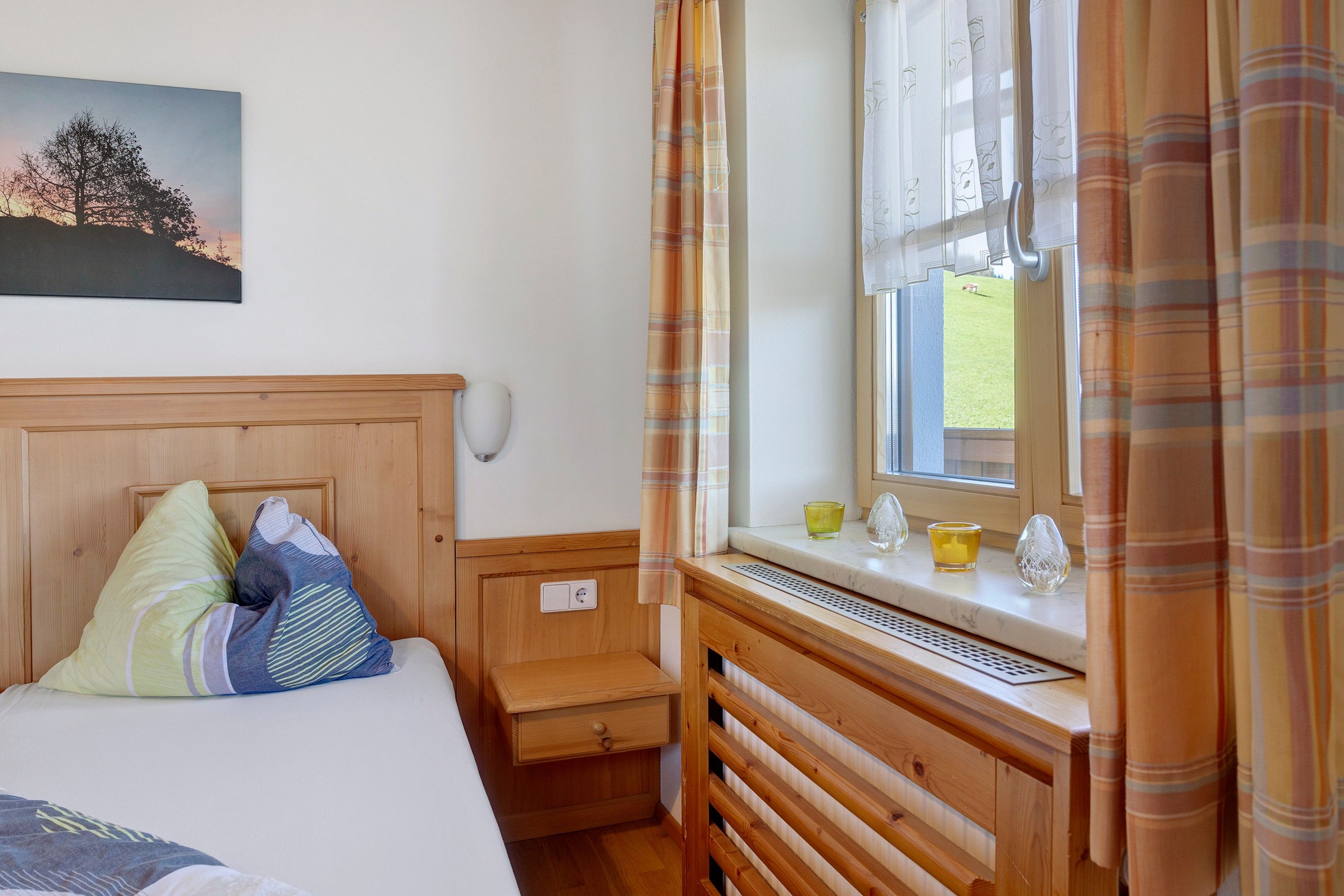 Ein Schlafzimmer mit hölzernem Kopfteil, einem Bett mit Kissen, einem Nachttisch, einem Fenster mit Vorhängen und Blick auf ein grünes Feld.