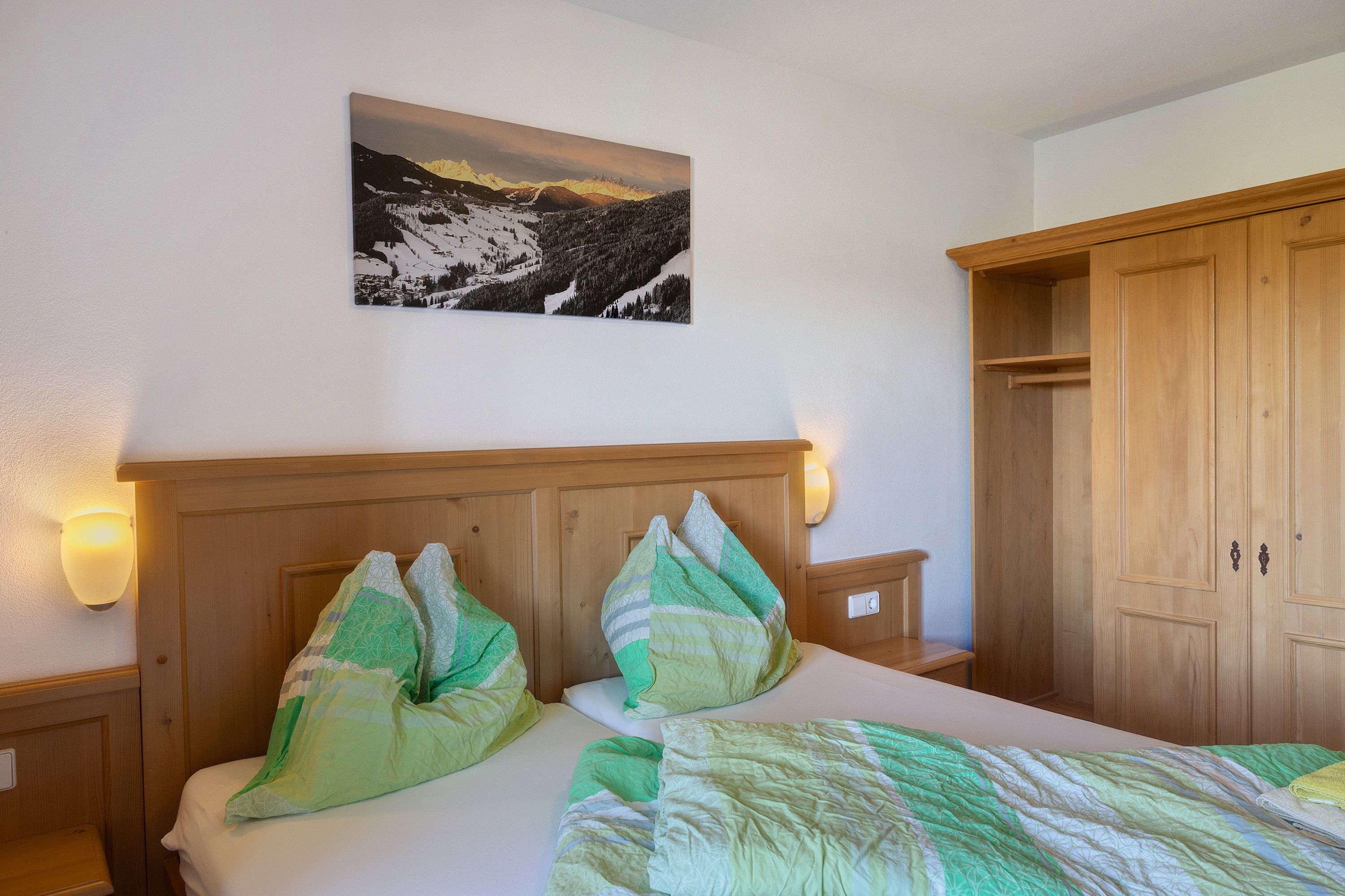 Un dormitorio con cabecera de madera, dos almohadas, cama con manta de diseño verde y blanco, un cuadro colgado en la pared de un paisaje montañoso y un armario de madera con puertas.