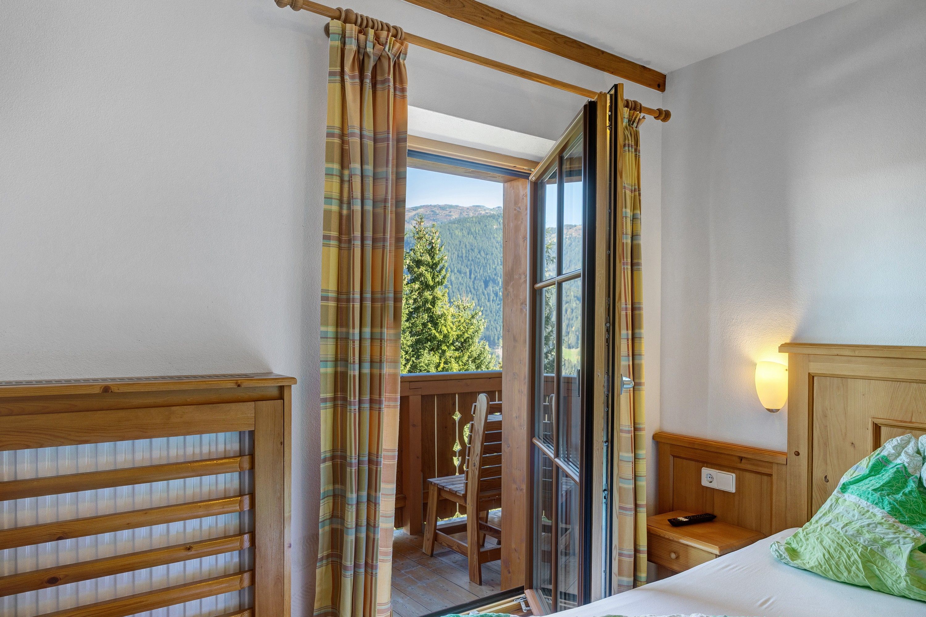 Una stanza con un letto, una testiera in legno, un comodino, una finestra con tende e vista su una catena montuosa ricoperta di boschi.