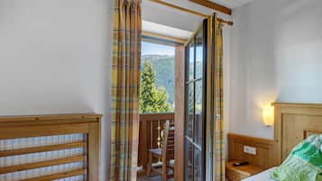 Una stanza con un letto, una testiera in legno, un comodino, una finestra con tende e vista su una catena montuosa ricoperta di boschi.