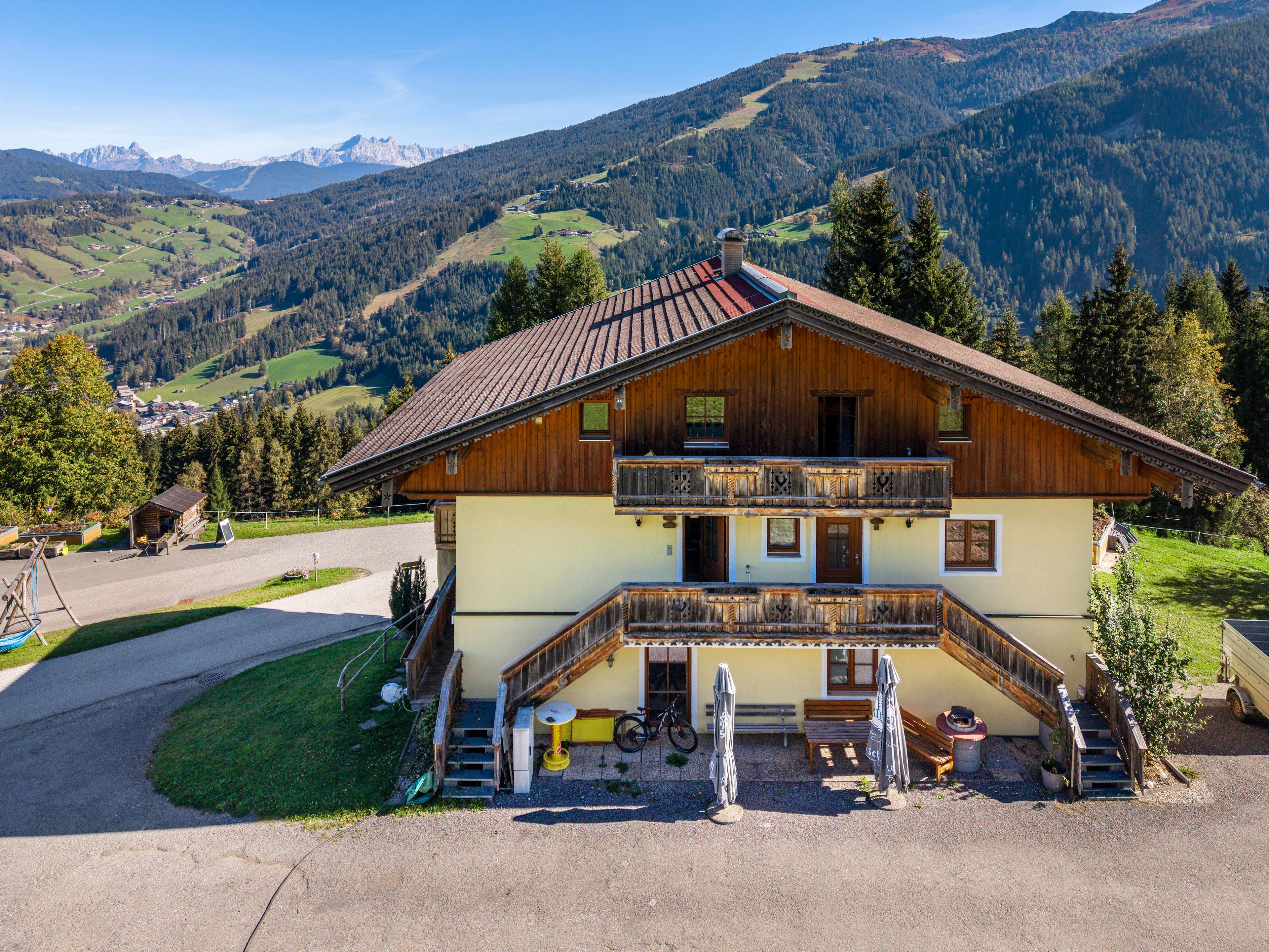 A two-story wooden house with a balcony, a driveway, and a view of a mountainous landscape.