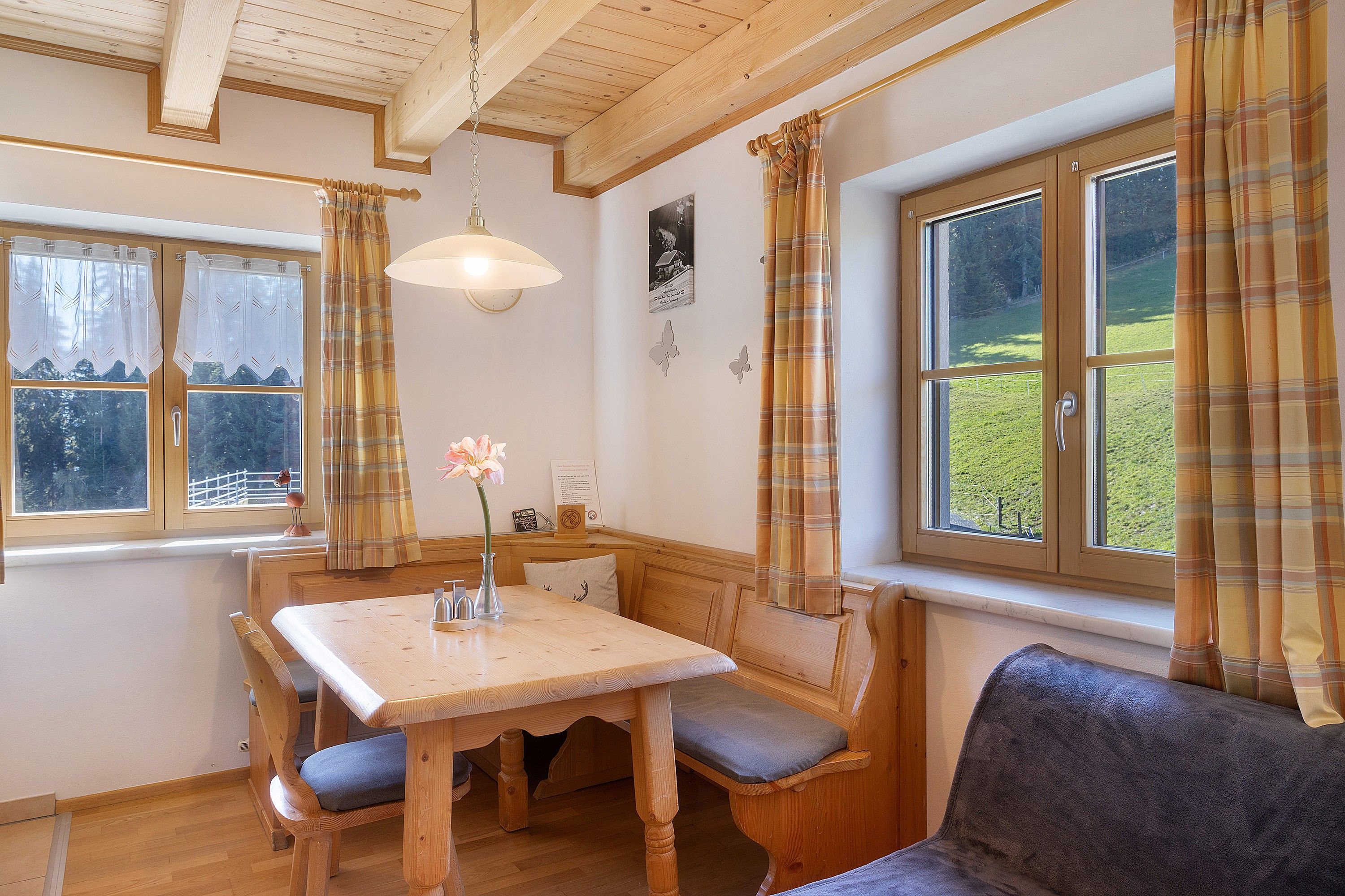 Un interior de madera con una mesa y bancos, una ventana con cortinas y una lámpara fijada a la pared.