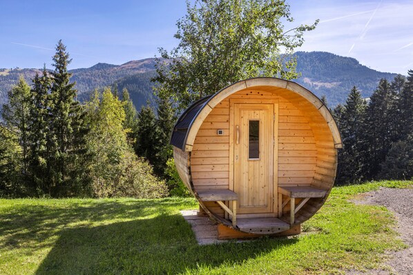 Een houten sauna met deur en treden, omgeven door gras en bomen, met bergen op de achtergrond.