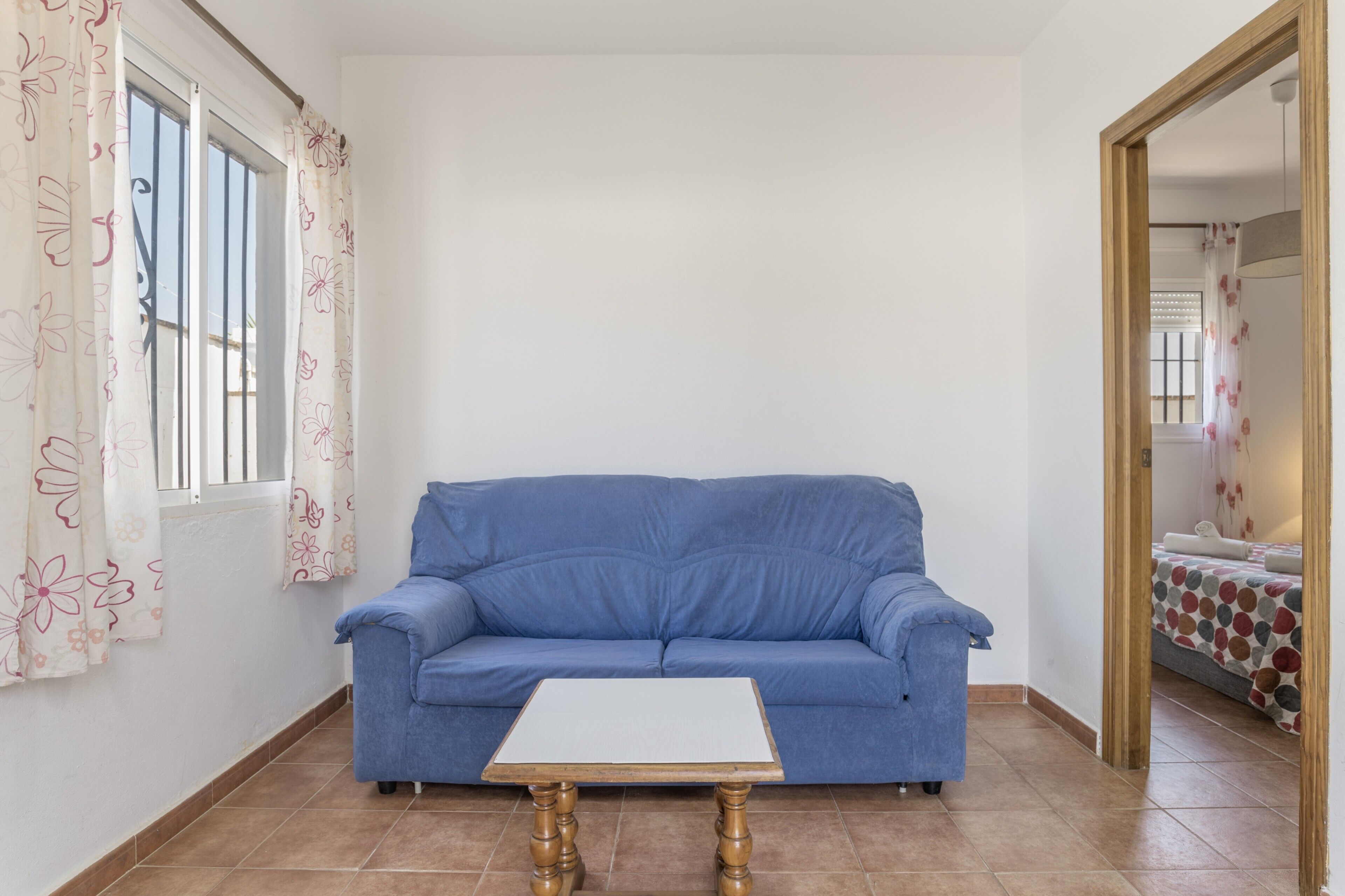Ein Zimmer mit einem blauen Sofa, einem weißen Tisch und einem Fenster mit blumigen Vorhängen.