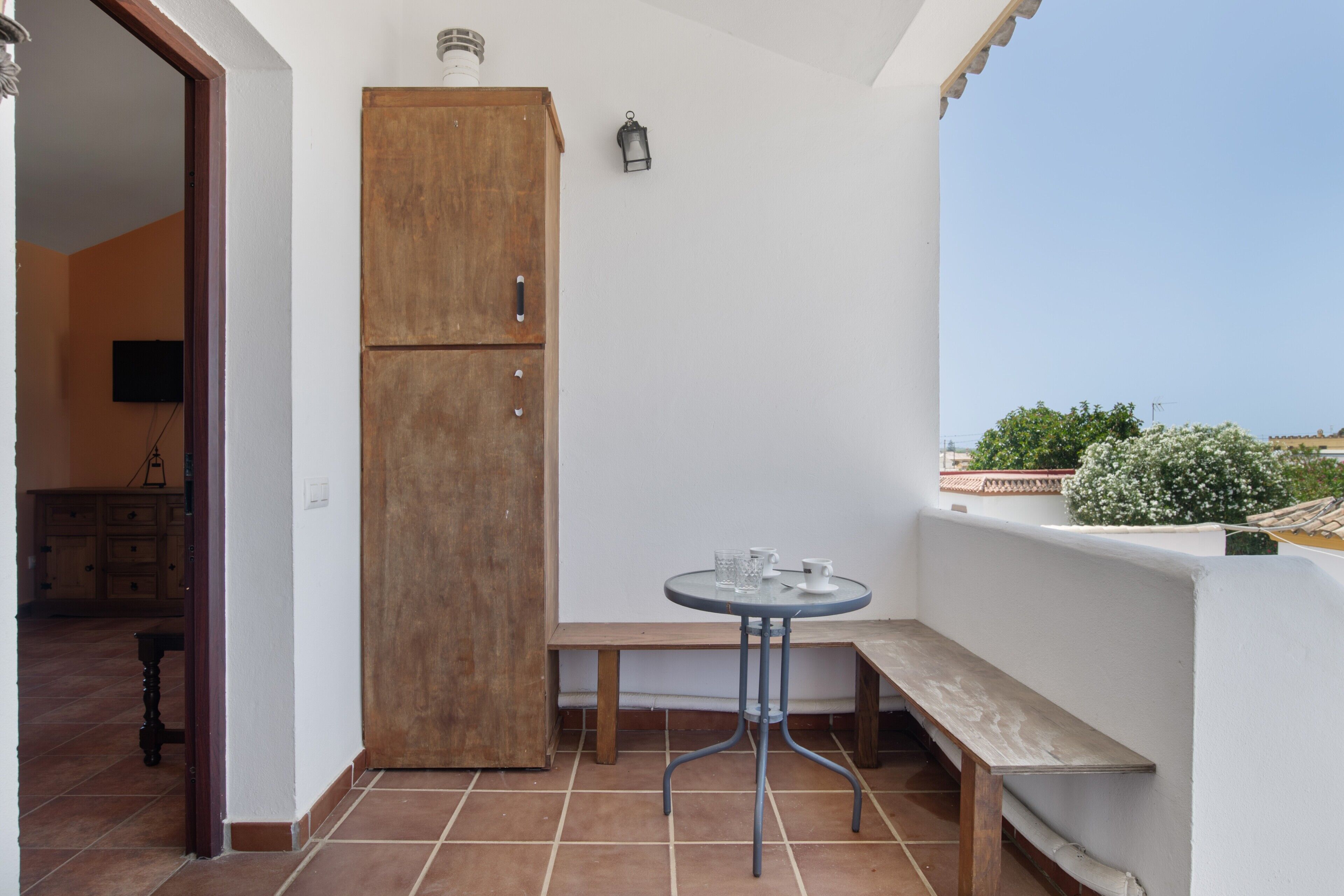 Un pequeño balcón con un mueble de madera, una mesa redonda con dos vasos, un banco y vistas de la ciudad.