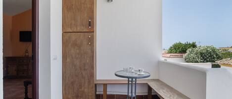 Un pequeño balcón con un mueble de madera, una mesa redonda con dos vasos, un banco y vistas de la ciudad.
