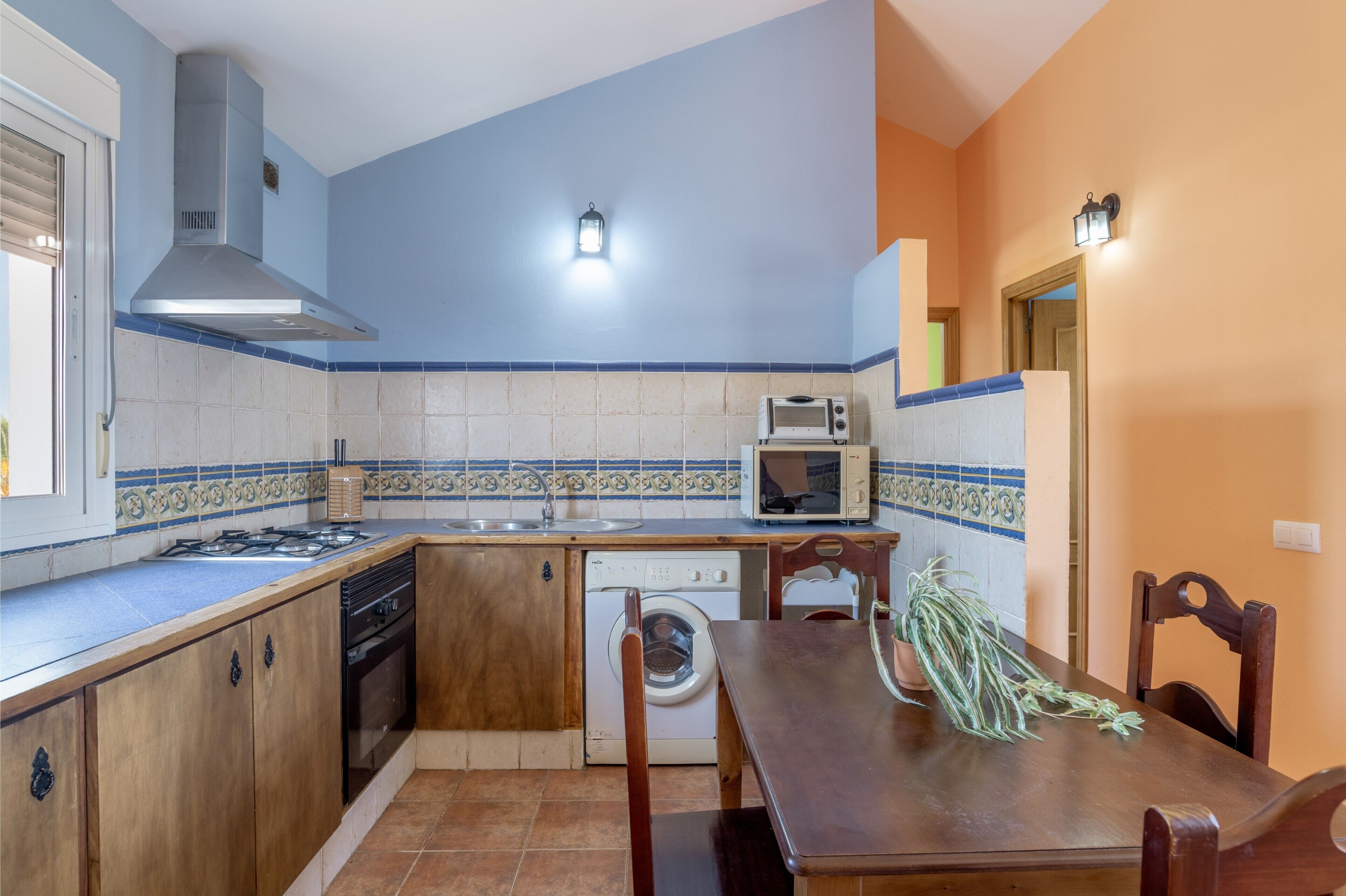 Une cuisine compacte équipée d’une cuisinière, d’un four à micro-ondes et d’un lave-linge, avec des meubles en bois et un coin repas comprenant des chaises.