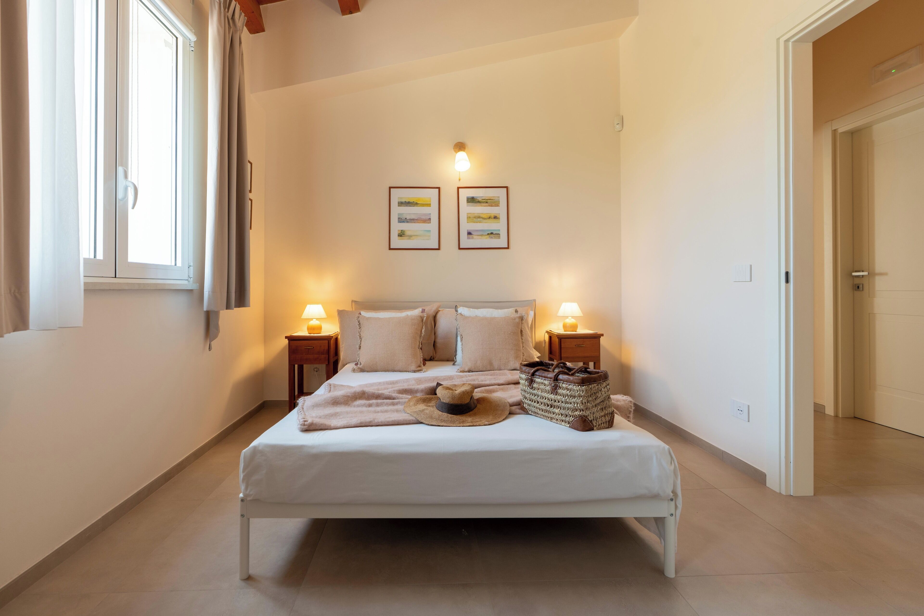 A neatly arranged bedroom with a bed, bedside lamps, a hat, and a woven bag.