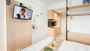 A hotel room with a bed, a TV mounted on the wall, a small kitchenette, and a shelf with a water bottle. - Comfy And Tidy Studio At Patraland Urbano Apartment (Bekasi)