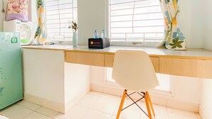 A room with a desk, two windows with curtains, a chair, a water dispenser, and a small table with a vase and bottles. - Comfy And Tidy Studio At Patraland Urbano Apartment (Bekasi)