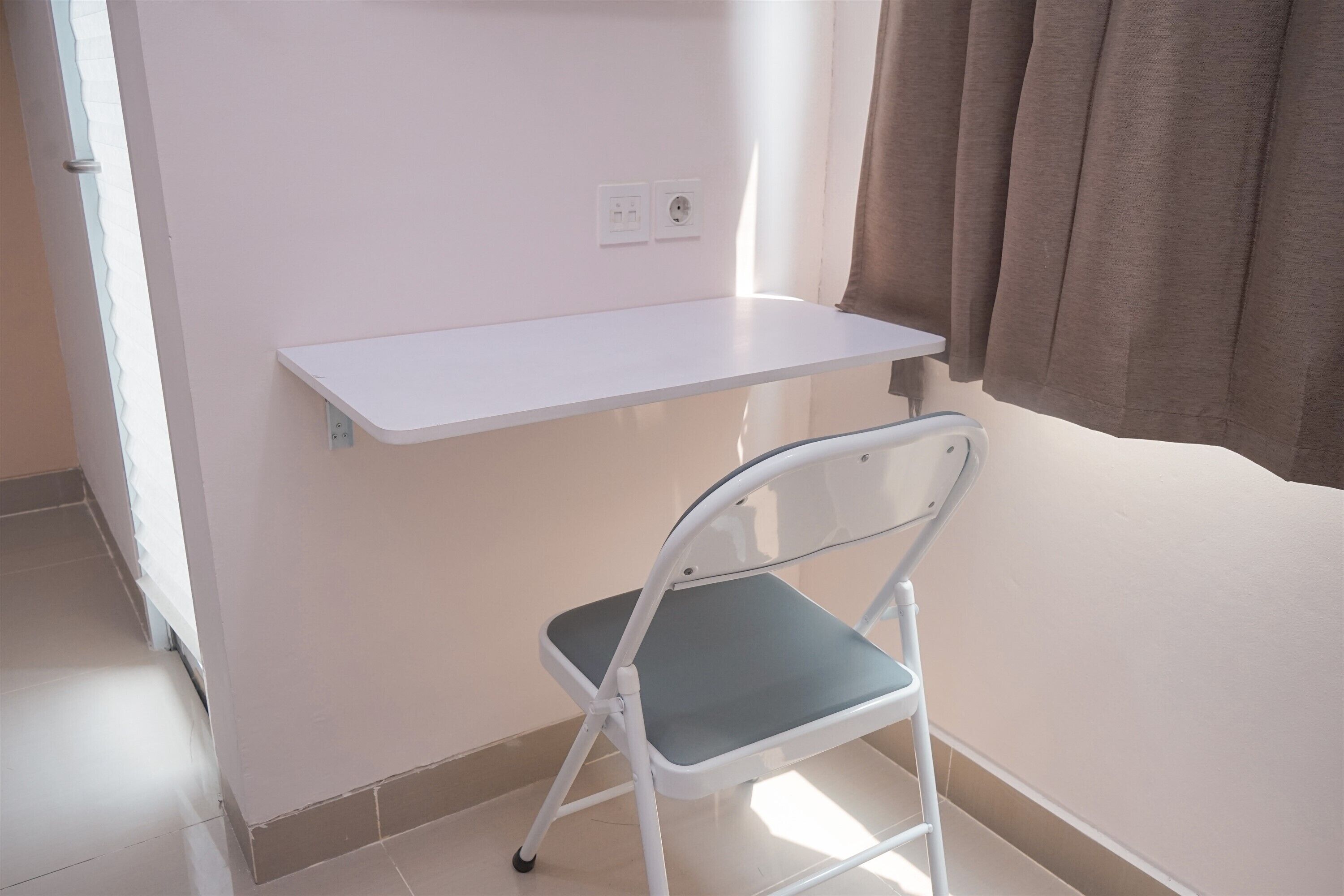 Una habitación pequeña con un escritorio blanco, una silla plegable y una ventana con cortinas.