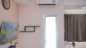 Ένα δωμάτιο ξενοδοχείου με ένα κρεβάτι, ένα γραφείο με έναν υπολογιστή, ένα κλιματιστικό τοποθετημένο στον τοίχο, μια πόρτα και ένα παράθυρο με κουρτίνες.
