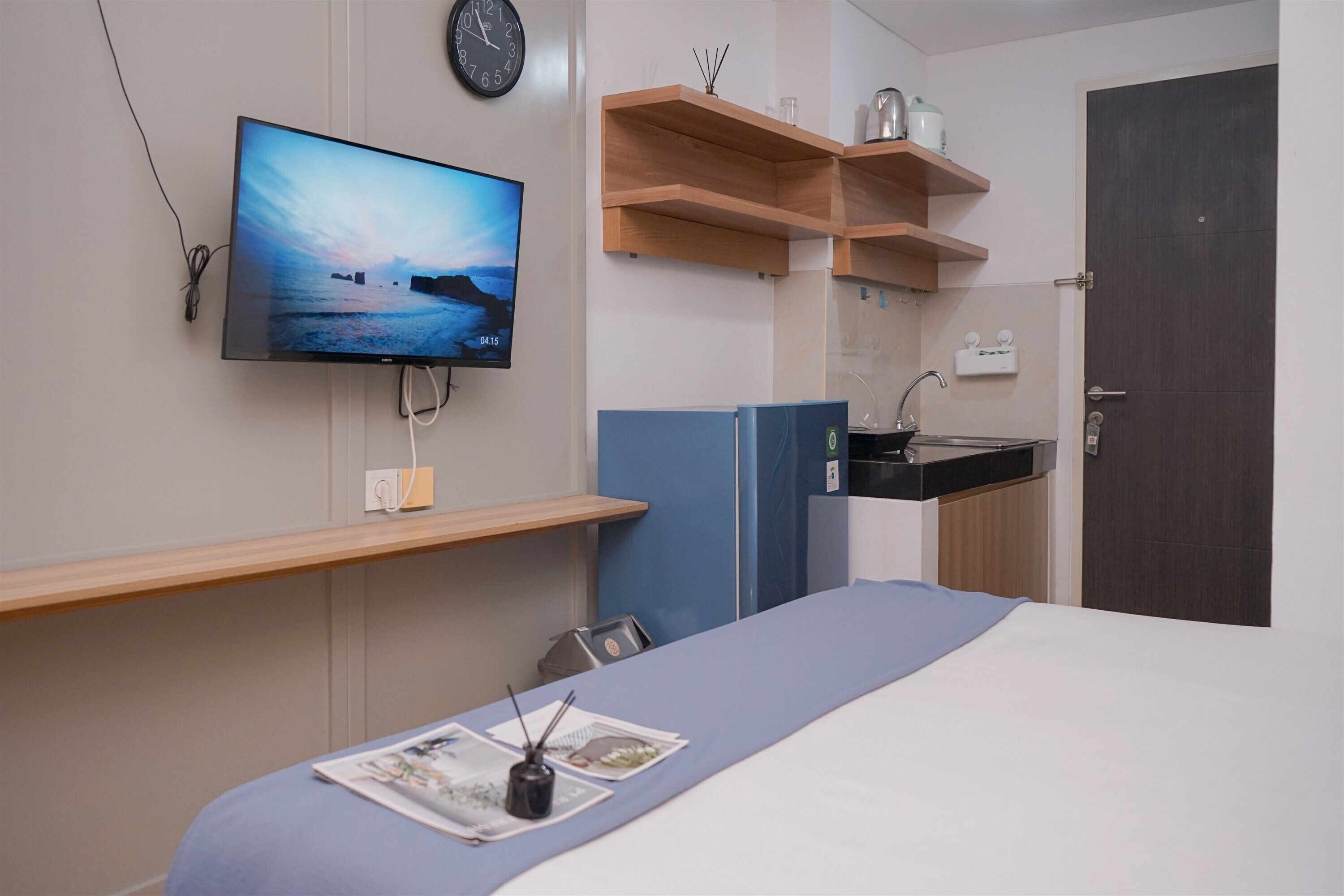 A hotel room with a bed, a TV mounted on the wall, a wooden shelf above the bed, a small kitchen area with a sink, and a door.