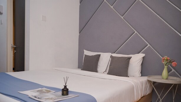 A neatly made bed with a blue blanket, a small table with a vase of flowers, and a wall with a geometric pattern. - Tidy And Restful Studio At Serpong Garden Apartment (Cisauk)