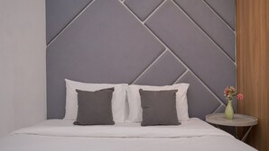 Una cama con ropa blanca y dos almohadas grises, una mesita redonda pequeña con un florero, y un cabecero de estampado geométrico.