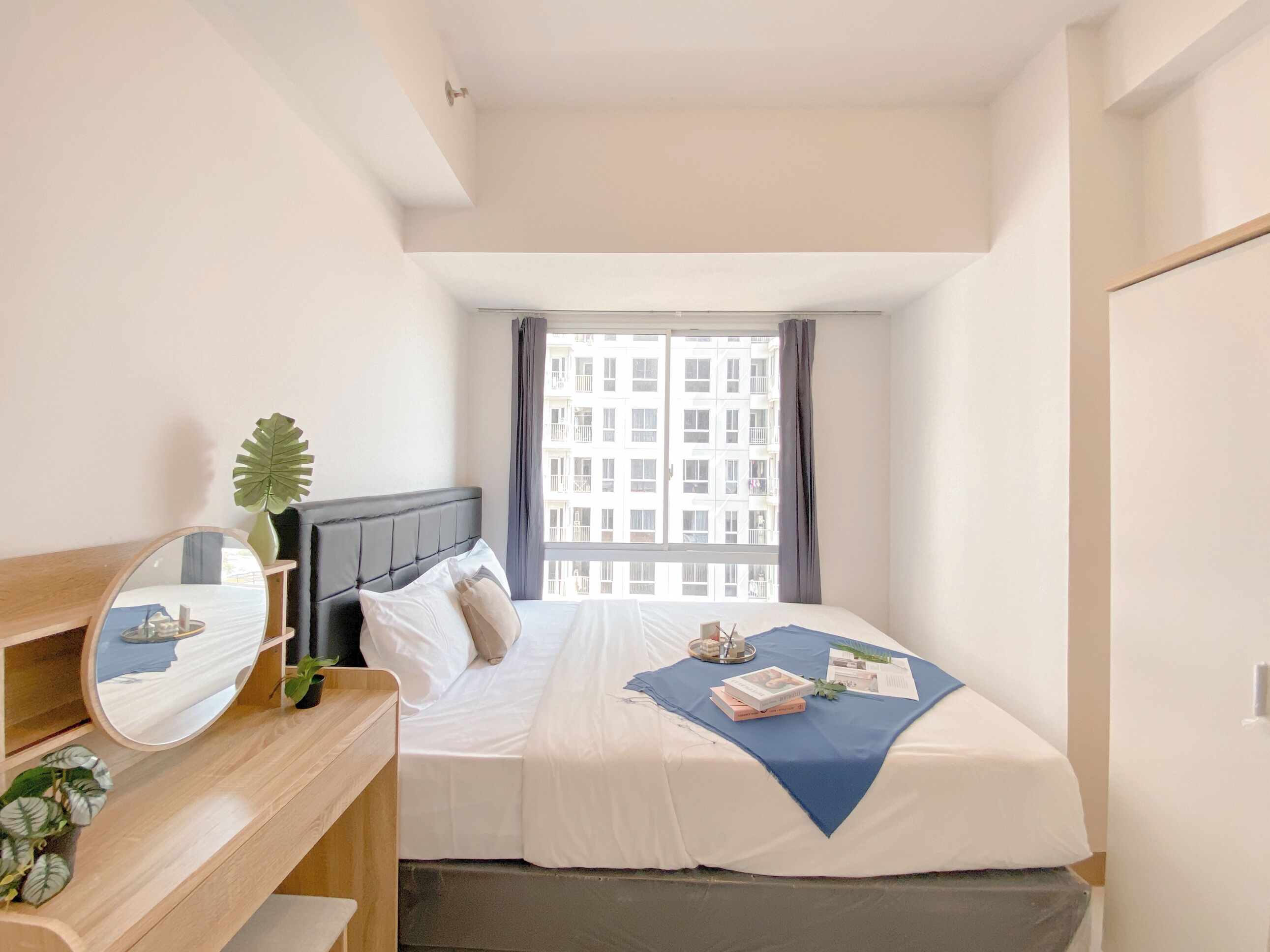 A hotel room with a bed, a bedside table with a mirror, a small plant, and a window with curtains.