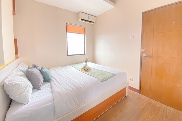 A bedroom with a bed, pillows, a bedside table with a vase, a window with blinds, an air conditioner, and a wooden door.