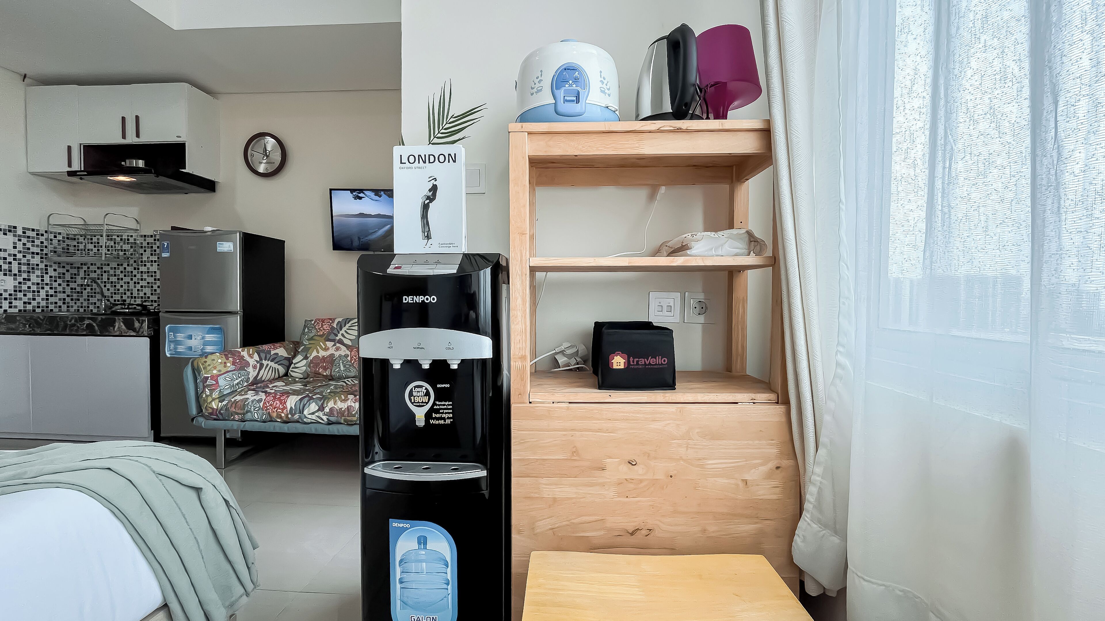 Una habitación de hotel moderna con kitchenette, dispensador de agua, repisa de madera con lámpara y cama con cojín estampado floral.