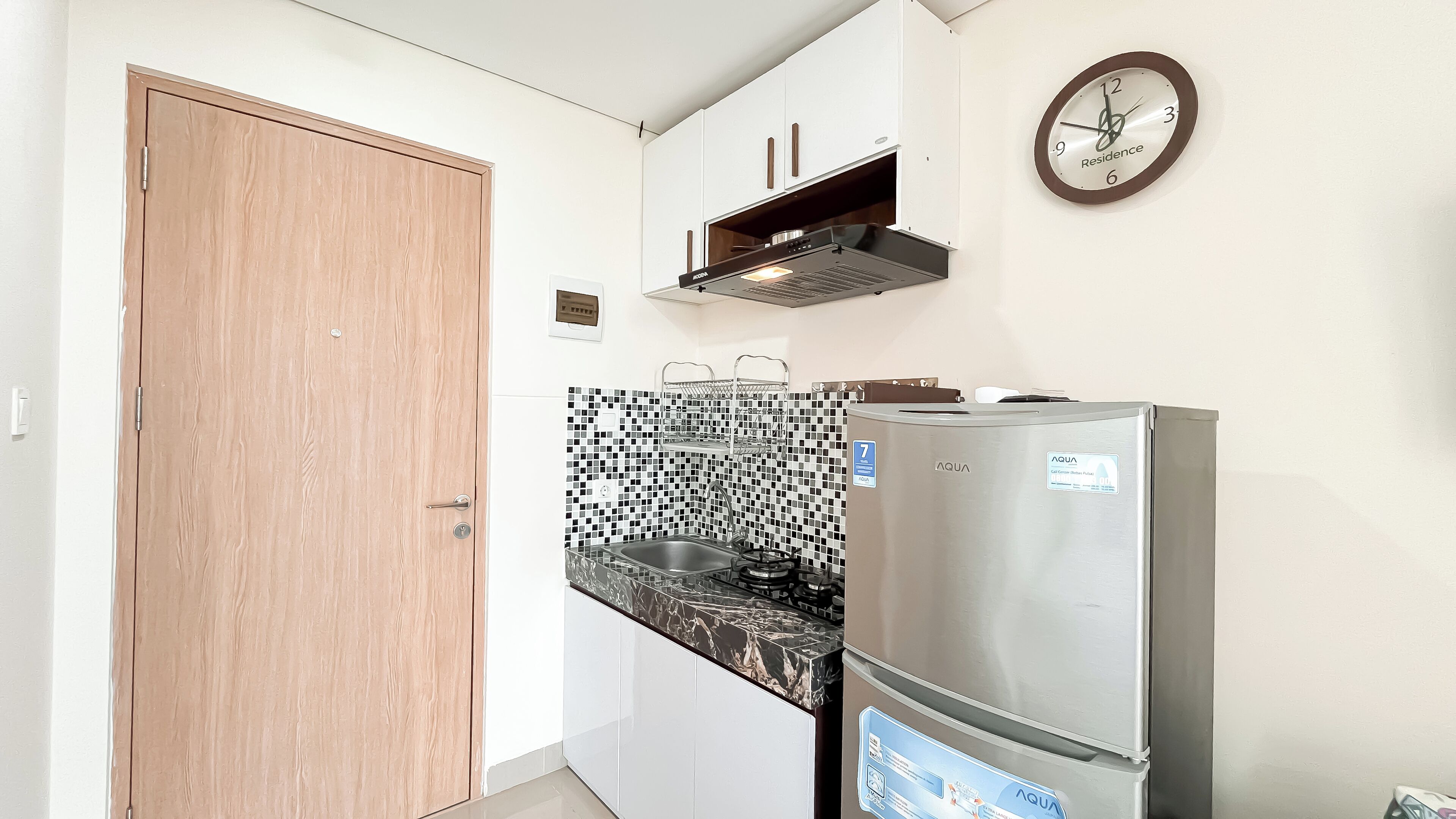Eine kleine Küche mit Holztür, Edelstahlkühlschrank, gefliestem Spritzschutz und einer Wanduhr.