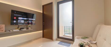 A modern living room with a flat-screen TV mounted on the wall, a sofa, a coffee table with decorative items, and a sliding glass door leading to a balcony.