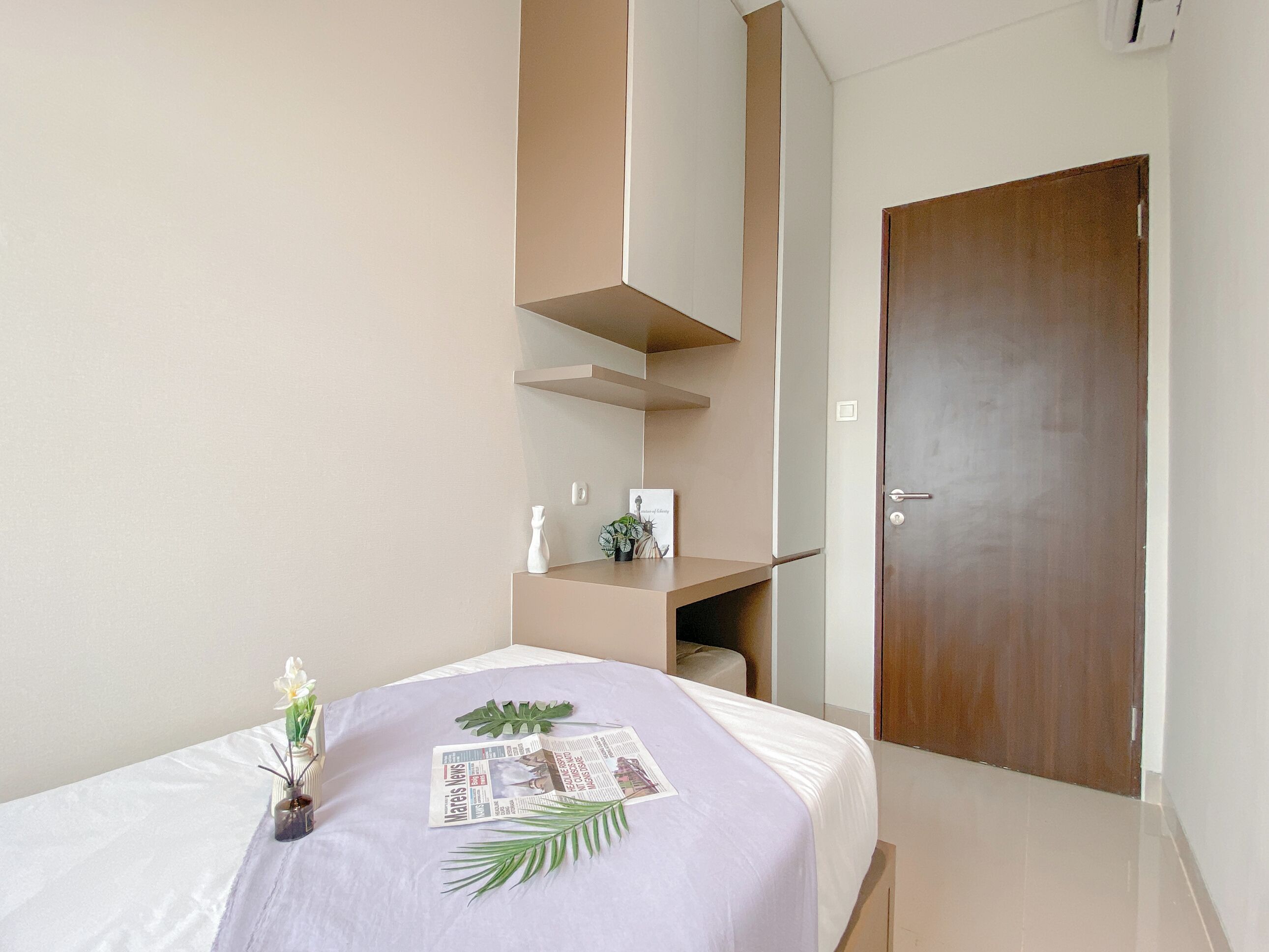 A hotel room with a bed, a small table with a vase and a plant, a wooden door, and a white wall.