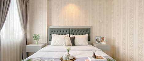 A neatly made bed with a tufted headboard, a bedside table with a plant, and a tray with books and a small vase.