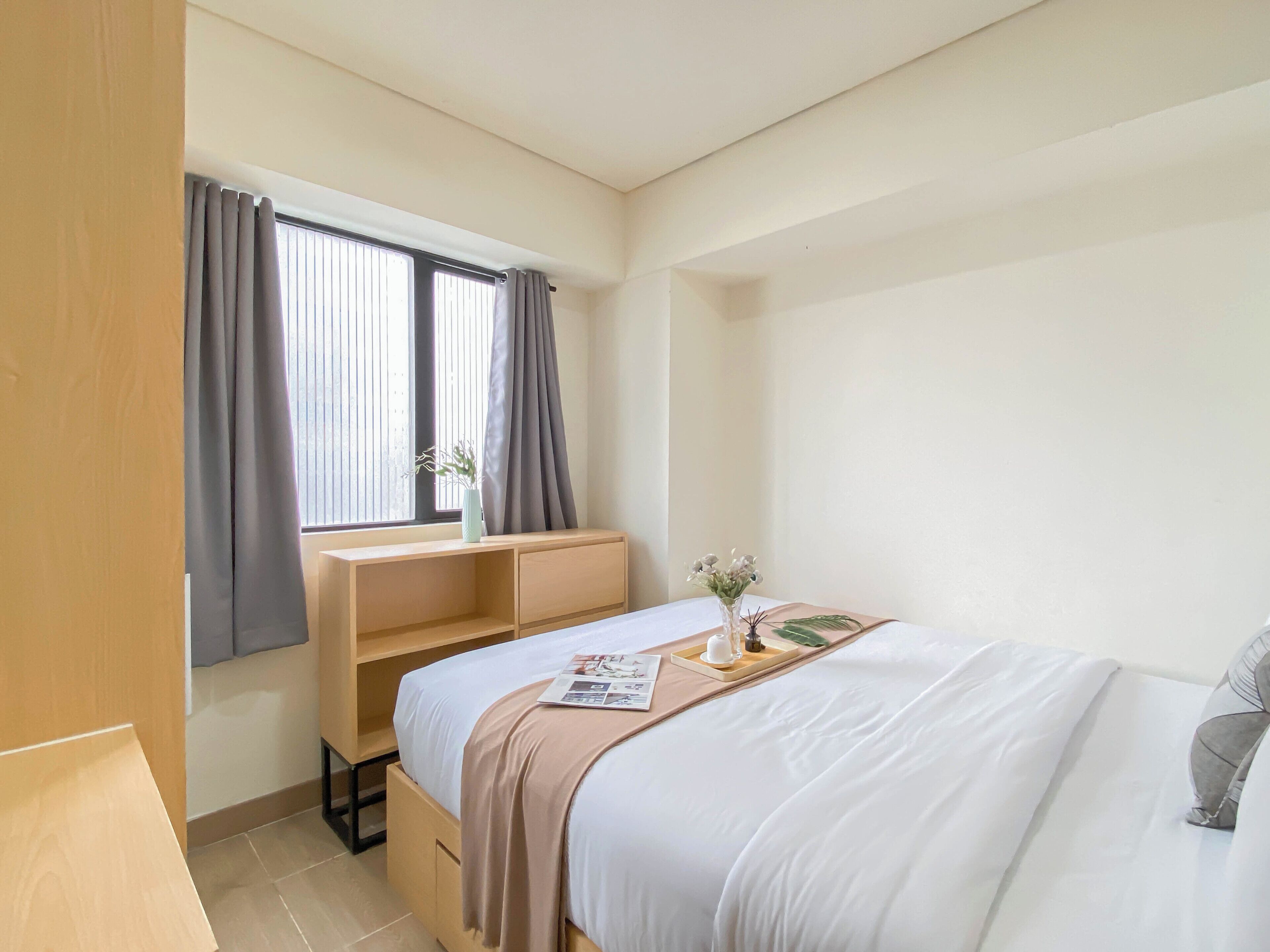 A hotel room with a bed, a wooden desk, a window with curtains, and a small table with a vase of flowers.
