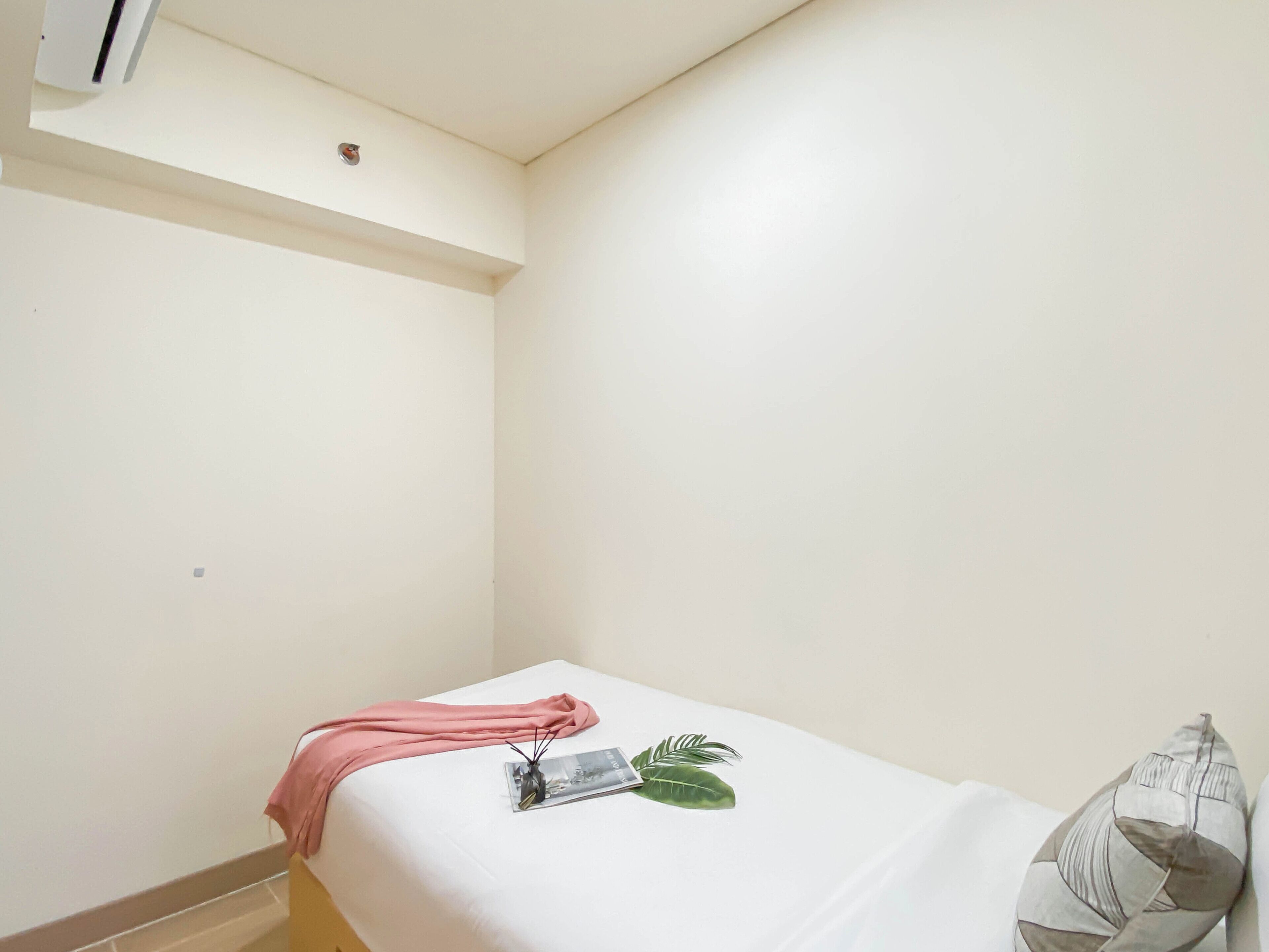 Ein kleines Zimmer mit einem Einzelbett, einer weißen Bettdecke, einem rosa Handtuch und einem Zierkissen.