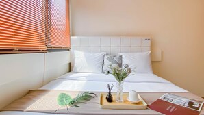 A neatly made bed with white bedding, a decorative pillow, a vase with flowers, a tray with a candle and a book, and a window with blinds.