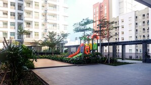 A playground with a slide and climbing structures, surrounded by greenery and modern buildings. - Warm And Homey 2Br Apartment At Green Bay Pluit (Jakarta)